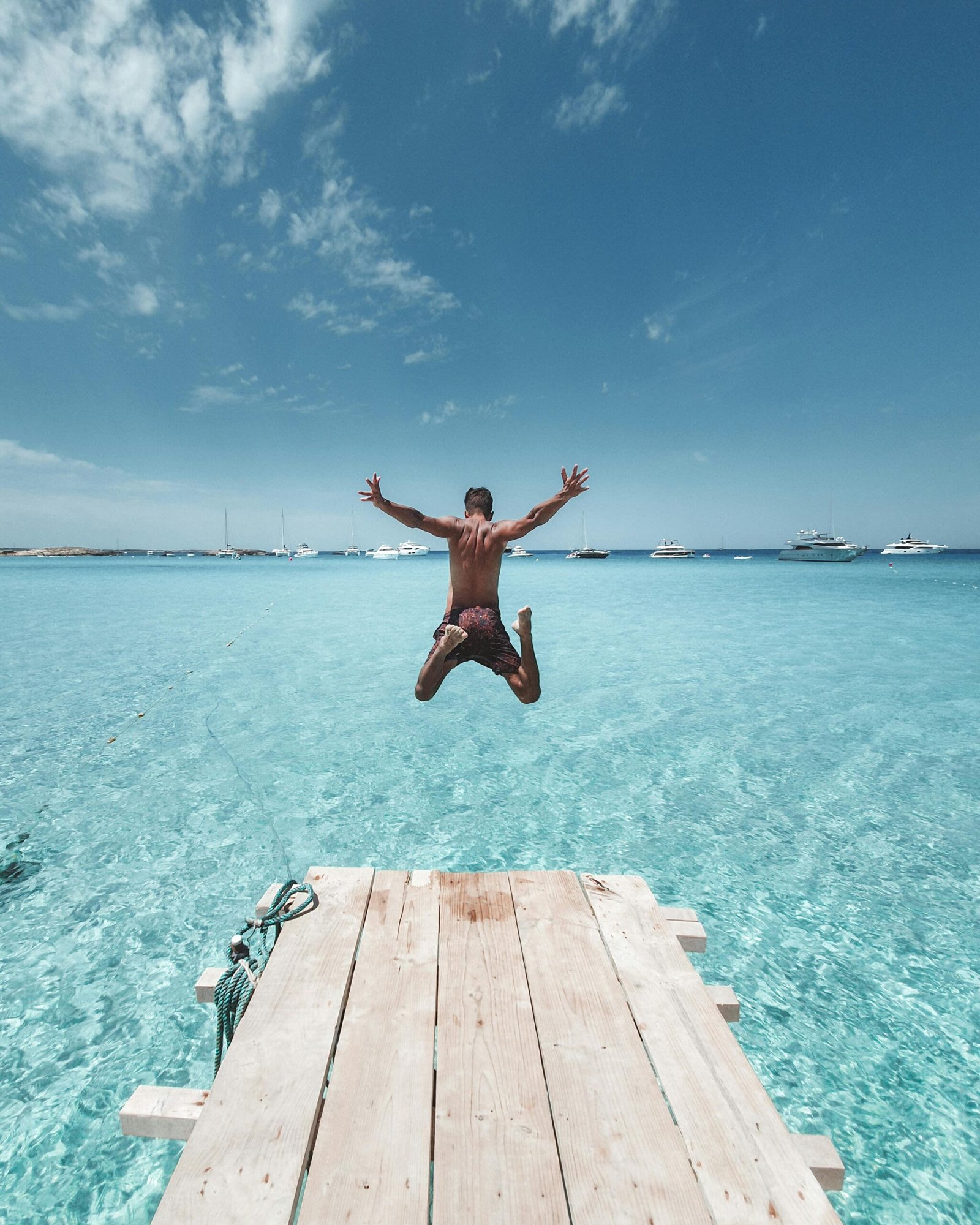 A man joyfully leaps off a wooden pier into the clear, turquoise waters on a sunny summer day.